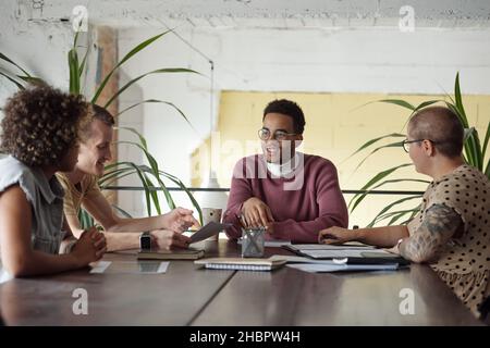 Group of young successful economists in casualwear sitting by table in office and discussing plans and ideas for new project Stock Photo
