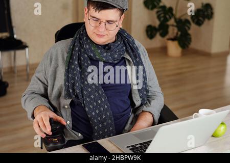 Man with disability rotating control panel of his wheelchair while sitting in front of laptop in domestic room Stock Photo