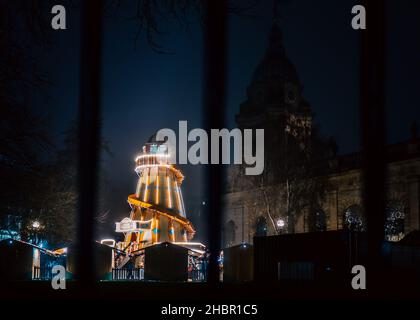Christmas helter skelter Christmas fairground rides lit up at night with Xmas lights and church behind. Low angle blue hour sky in the evening. Stock Photo