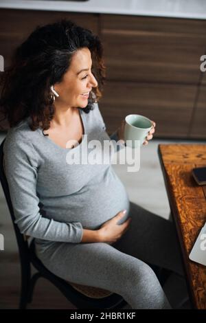 Pregnant female freelancer talking through bluetooth while having coffee at home Stock Photo
