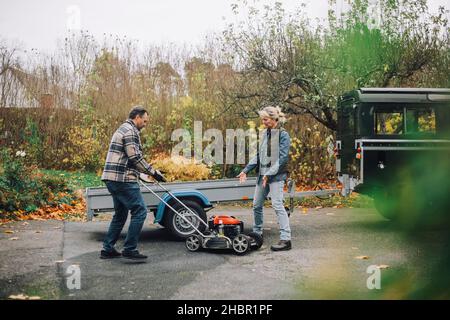 Mature man holding lawn mower while talking with female friend on driveway Stock Photo