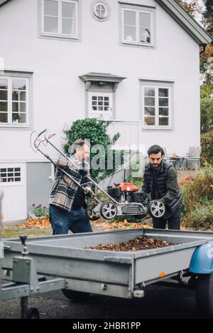 Son and father loading lawn mower in vehicle trailer by building Stock Photo