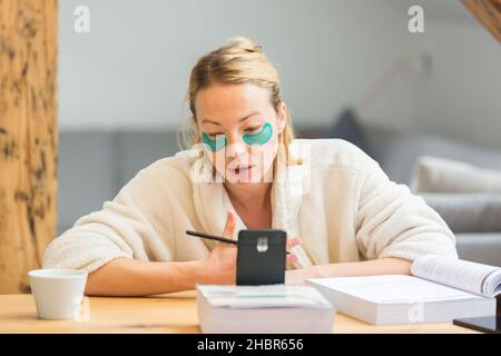 Young businesswoman wearing cosy warm bathrobe having remote cell phone conference call meeting from home living room in winter Christmas time. Remote Stock Photo