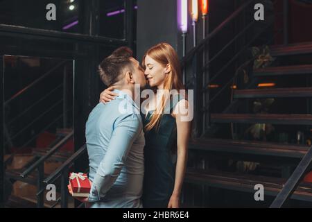 Man hiding behind his back a gift box of while kissing girlfriend. Happy birthday holiday. Dating Stock Photo