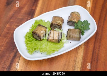 Eggplant rolls decorated with fresh lettuce and parsley on white plate, wooden background. Top view, close up with copy space, Georgian cuisine.  Stock Photo