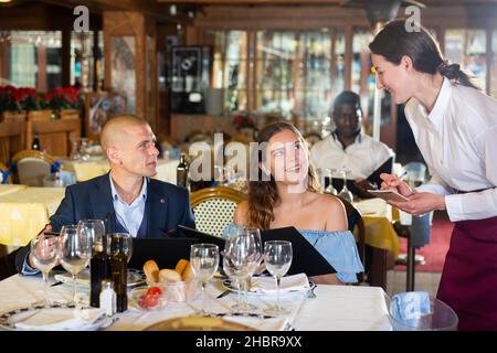 Attractive smiling woman waiter receiving order from guest in fashionable restaurant Stock Photo