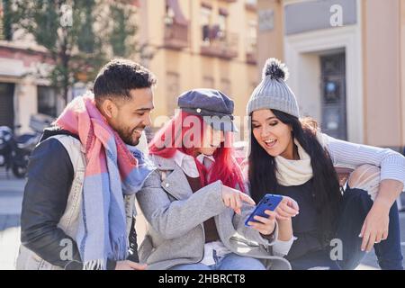 Stylish female with long red hair sharing smartphone with multiracial friends and looking at screen together on city street Stock Photo