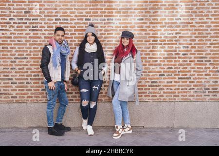 Positive multiracial young male and female friends wearing trendy casual outfits standing near brick wall and smiling at camera Stock Photo