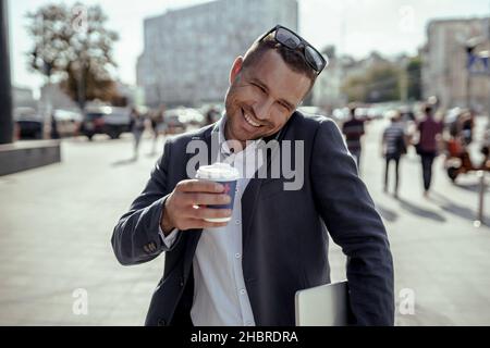 Young businessman holding his laptop while using his phone and drinking coffee. Standing on the street. Stock Photo