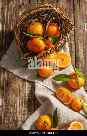 Tangerines falling out of the basket. Oranges, mandarins, clementines, citrus fruits) with green leaves. Wooden background, rustic style, top view Stock Photo