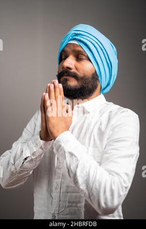side view shot of indian sikh man doing namste or praying to god on gray background with closed eyes - conept of faith, religious beliefs and Sikhism Stock Photo