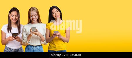 Three Young Women Using Smartphones While Standing Isolated Over Yellow Studio Background Stock Photo