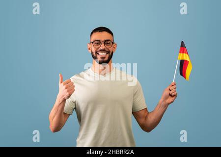 Happy arab guy showing thumb up and flag of Germany, posing over blue studio background. Education in Germany Stock Photo