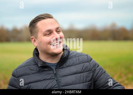 Cheerful upbeat young man in a warm winter jacket standing looking aside with a happy beaming smile outdoors in a park Stock Photo