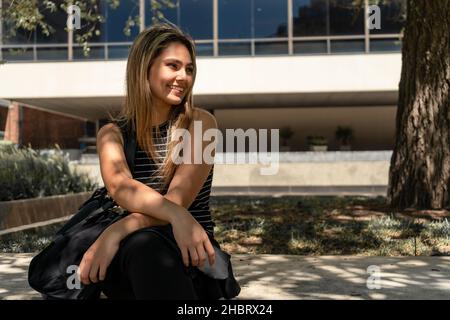 A portrait of a beautiful young smiling woman sitting on a bench in the shade of a tree with a modern building in the background. Stock Photo