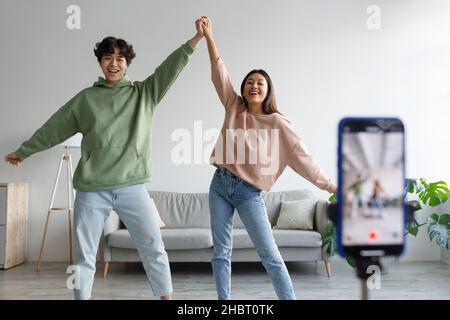 Happy young Asian couple making video on cellphone, dancing in front of camera at home Stock Photo