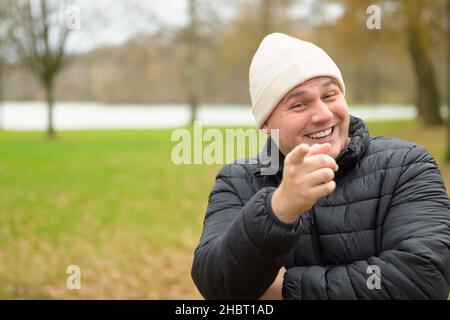 Laughing young man in warm winter jacket and beanie pointing a finger at the camera outdoors in a park Stock Photo