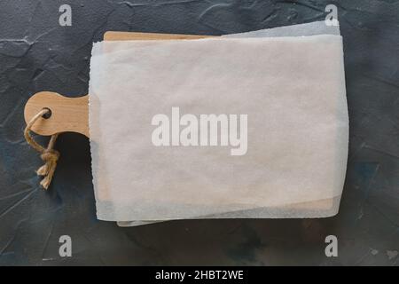 Wooden cutting board and white parchment paper on dark gray background. Mockup for recipes, shopping list or food background. Top view, copy space Stock Photo