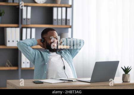 Happy young black bearded man in glasses and headphones rejoices in successful deal, relax and looks at laptop Stock Photo