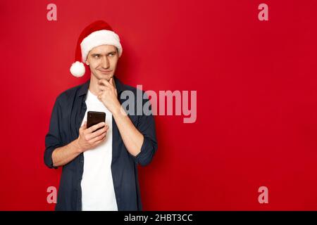 young man with phone in hand smiles slyly, thinks about new idea congratulations Christmas Santa hat Stock Photo