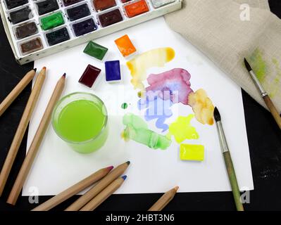 Artistic paints, a glass of water, pencils and a sheet of paper. Watercolor paints in a plastic container and colored paint strokes on a sheet. Brush Stock Photo
