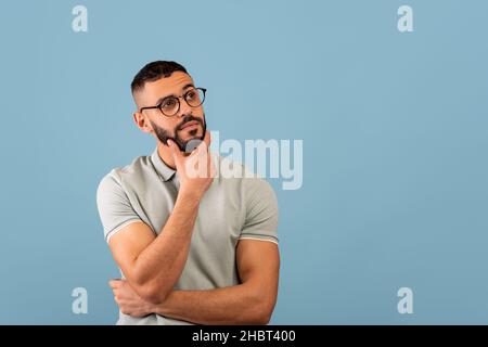 Pensive middle eastern guy thinking and touching chin, looking aside at free space, posing over blue background Stock Photo