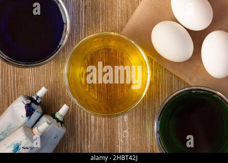 Top view of three bowls with colored water for colouring eggs in blue, yellow, green color and white eggs on wooden background and food coloring tubes Stock Photo