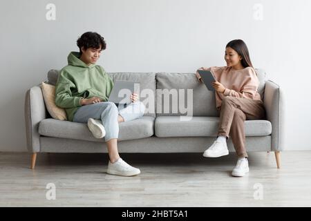 Handsome young Asian guy and his girlfriend using modern gadgets while sitting on couch at home, copy space Stock Photo