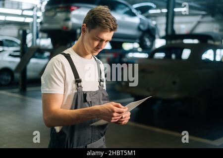 Side view of focused handsome young mechanic male wearing uniform reading clipboard standing in auto repair shop garage Stock Photo