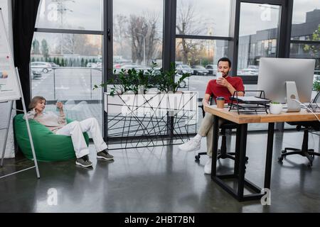 blonde woman sitting in bean bag chair and using smartphone near african american colleague in office Stock Photo