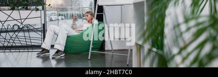 blonde manager sitting in bean bag chair and using smartphone, banner Stock Photo