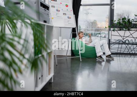 blonde manager sitting in bean bag chair and using smartphone Stock Photo