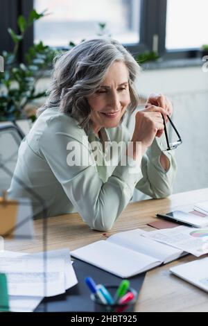 Cheerful businesswoman holding eyeglasses near documents and gadgets on table in office Stock Photo