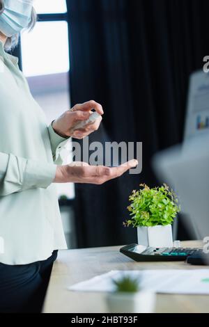 Cropped view of mature businesswoman in protective mask using hand sanitizer near working table in office Stock Photo