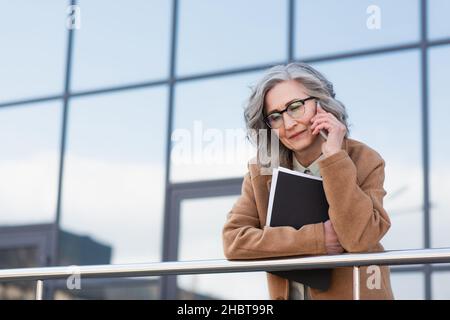 Upset businesswoman in coat talking on smartphone and holding papers on urban street Stock Photo