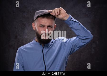 Young bearded european guy shows serious emotions being surprised by taking up the visor of a fashionable cap. Portrait on a dark background Stock Photo