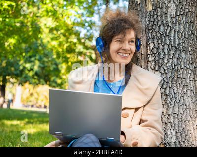 Smiling middle aged woman in headphones with laptop in city park. Close-up photo. Stock Photo