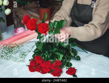Woman florist creating a bouquet of fresh red roses. Cropped image of female florist arranging rose flowers using tools. Concept of working with flowers, floral business. Selective focus Stock Photo