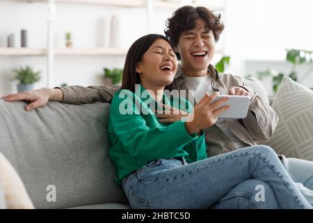 Joyful young Asian couple watching funny videos on smartphone, laughing together, relaxing on couch at home Stock Photo