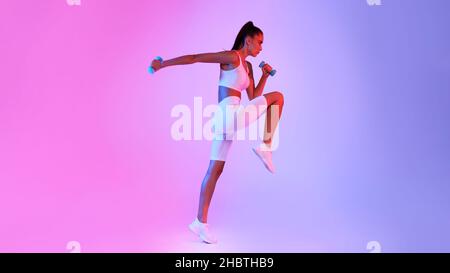Determined Fitness Lady Jumping Exercising Holding Dumbbells Over Neon Background Stock Photo