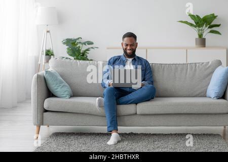 Happy young black bearded male chatting in social network, businessman work on laptop on sofa Stock Photo