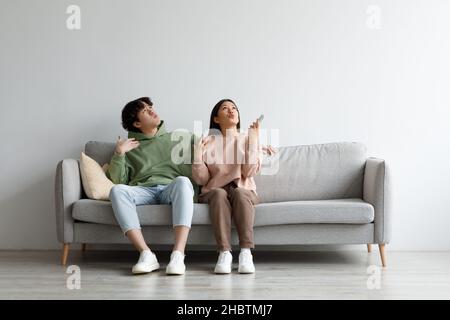 Young Asian couple suffering from summer heat, holding remote control, switching on air conditioner at home, mockup Stock Photo