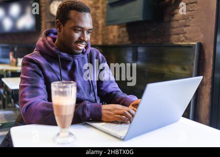 Busy African American male teleworker sitting at table with glass of chocolate milkshake and typing on netbook keyboard while working online in cafe Stock Photo