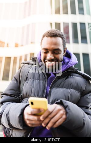 Positive black male in outerwear smiling and reading messages on smartphone while standing against contemporary skyscraper in city Stock Photo