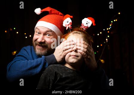 Dad in a New Year's hat makes a surprise to his son, covering his eyes with his hands. Stock Photo