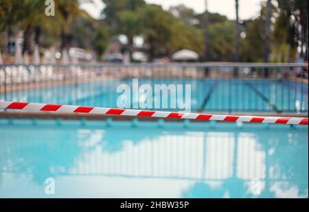 Red white tape in front of empty hotel resort swimming pool closed during evening cleaning, blurred water background Stock Photo