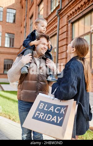 Vertical portrait of happy family holding shopping bags with Black Friday sign outdoors in autumn Stock Photo