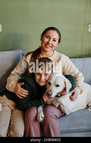 Vertical portrait of happy mother embracing son and dog in cozy home interior against green wall Stock Photo