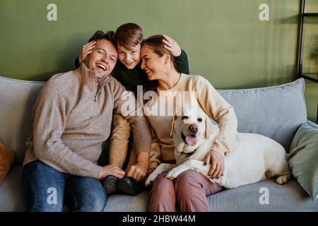 Portrait of happy family embracing son and dog while sitting on couch at home against green wall, copy space Stock Photo