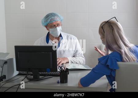 A doctor in a white coat, mask and cap in the office sits at a table with a patient and listens to her complaints. Consultative reception . Selective Stock Photo